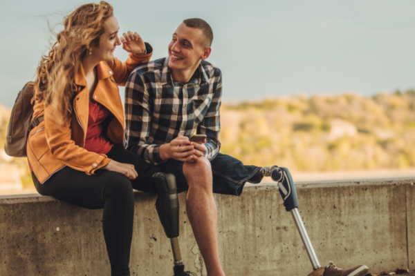 A woman with a prosthesis and a man with a prosthesis sitting on a small wall looking at each other and smiling.