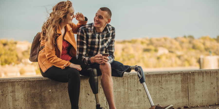 A woman with a prosthesis and a man with a prosthesis sitting on a small wall looking at each other and smiling.