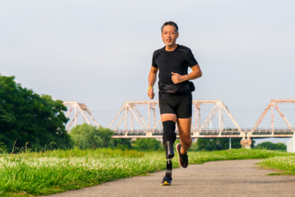 A man in a black shirt, black shorts, and wearing a prosthesis running on a sidewalk with grass on both sides. A bridge is in the background.